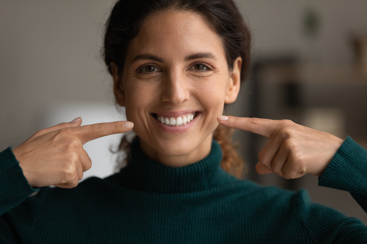 Head shot close up sincere attractive young hispanic latin woman pointing fingers at toothy smile, feeling satisfied with professional dental whitening procedure, showing healthy straight teeth.