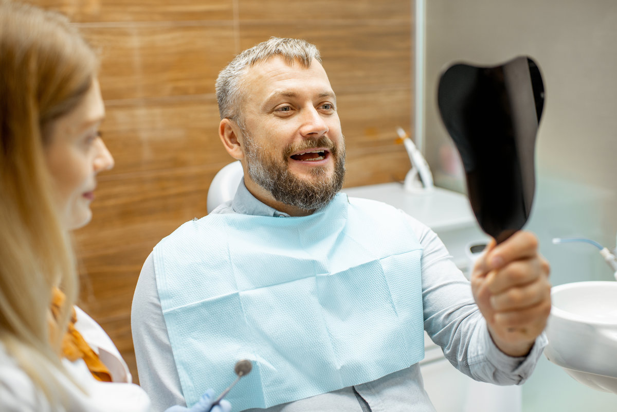 Man looking on the dental mirror, satisfied with his teeth, during a medical consultation with female dentist at the dental office