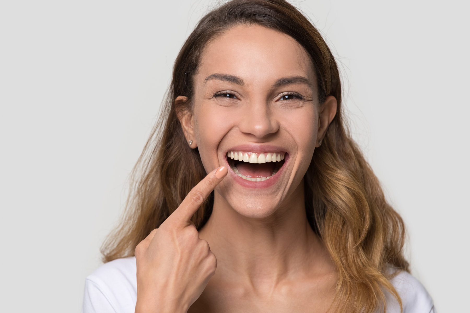 Happy young woman with white straight teeth perfect dent orthodontic smile pointing at tooth looking at camera isolated on studio blank background, dental health stomatology service concept, portrait